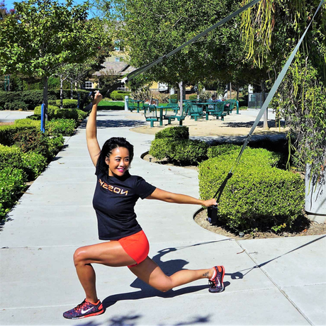 Fit woman working out in a part on a NOSSK Suspension Fitness Strap doing a suspended lunge as she smiles happily